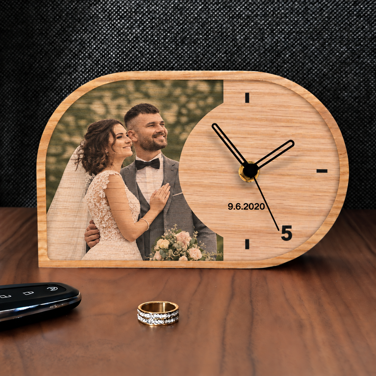 A personalized wooden clock with an arched top edge, featuring a photo of a bride and groom engraved on the left side and a light wood grain face on the right. The clock is shown on a wooden table next to wedding rings and a car key, with a dark textured background.