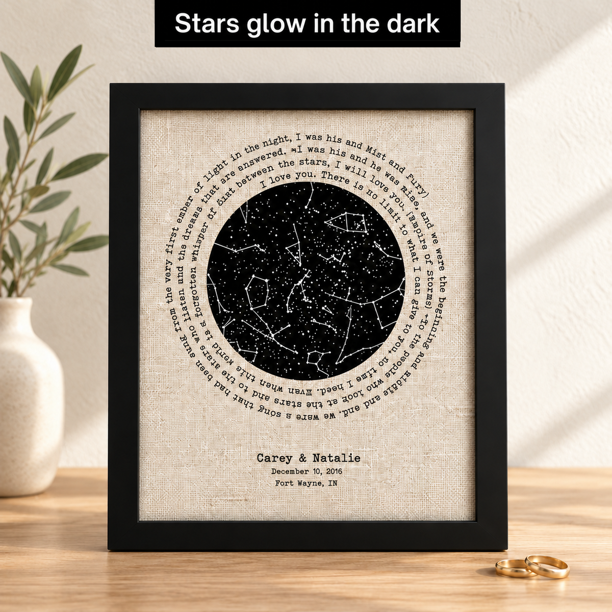A framed personalized constellation print with a black frame and textured beige background, featuring a circular star map and custom text for 'Carey & Natalie' on a wooden surface. A black banner at the top reads 'Stars glow in the dark', with a plant on the left and gold rings on the right.