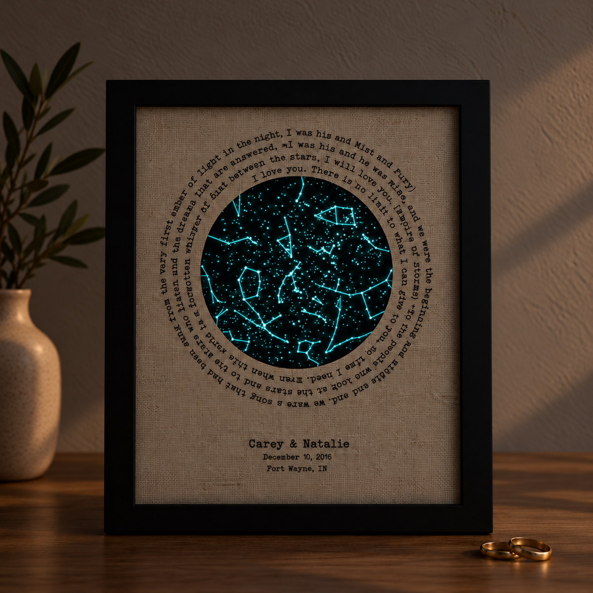A framed personalized constellation art print displayed on a wooden surface. The print features a black circular frame containing a textured beige background with a glowing blue constellation design in the center, surrounded by printed love quotes, and customized names 'Carey & Natalie' with a date and location at the bottom.